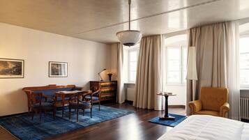 A hotel room with a wooden floor, a bed, a sofa, a desk, and a chair. There is a window with curtains and a lamp on a side table.