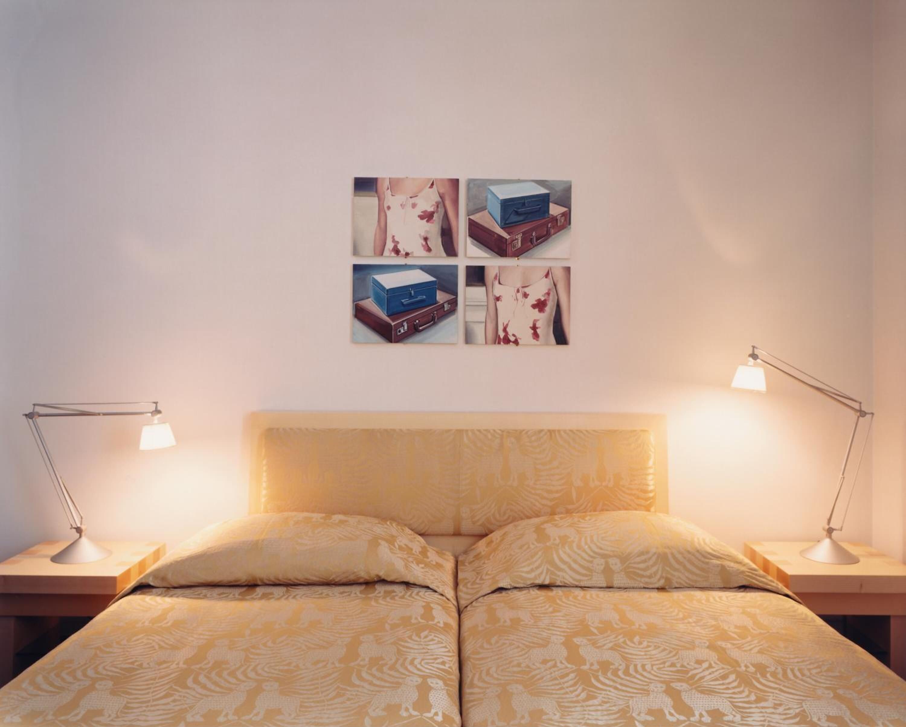 Una cama doble con un edredón estampado, dos mesitas de noche con lámparas, y una pared con cuatro cuadros de maletas.