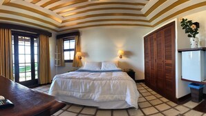 A hotel room with a bed, a desk with a phone, a lamp, a wardrobe, and a refrigerator.
