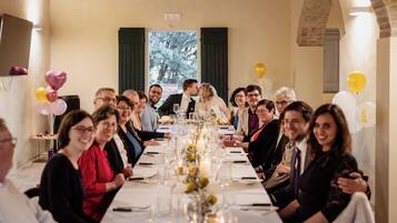 A long dining table with guests seated, a chandelier, and a view of the outdoors.