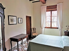 Una habitación con una cama, un escritorio de madera, una silla, un ventanal con cortinas y dos cuadros en la pared.