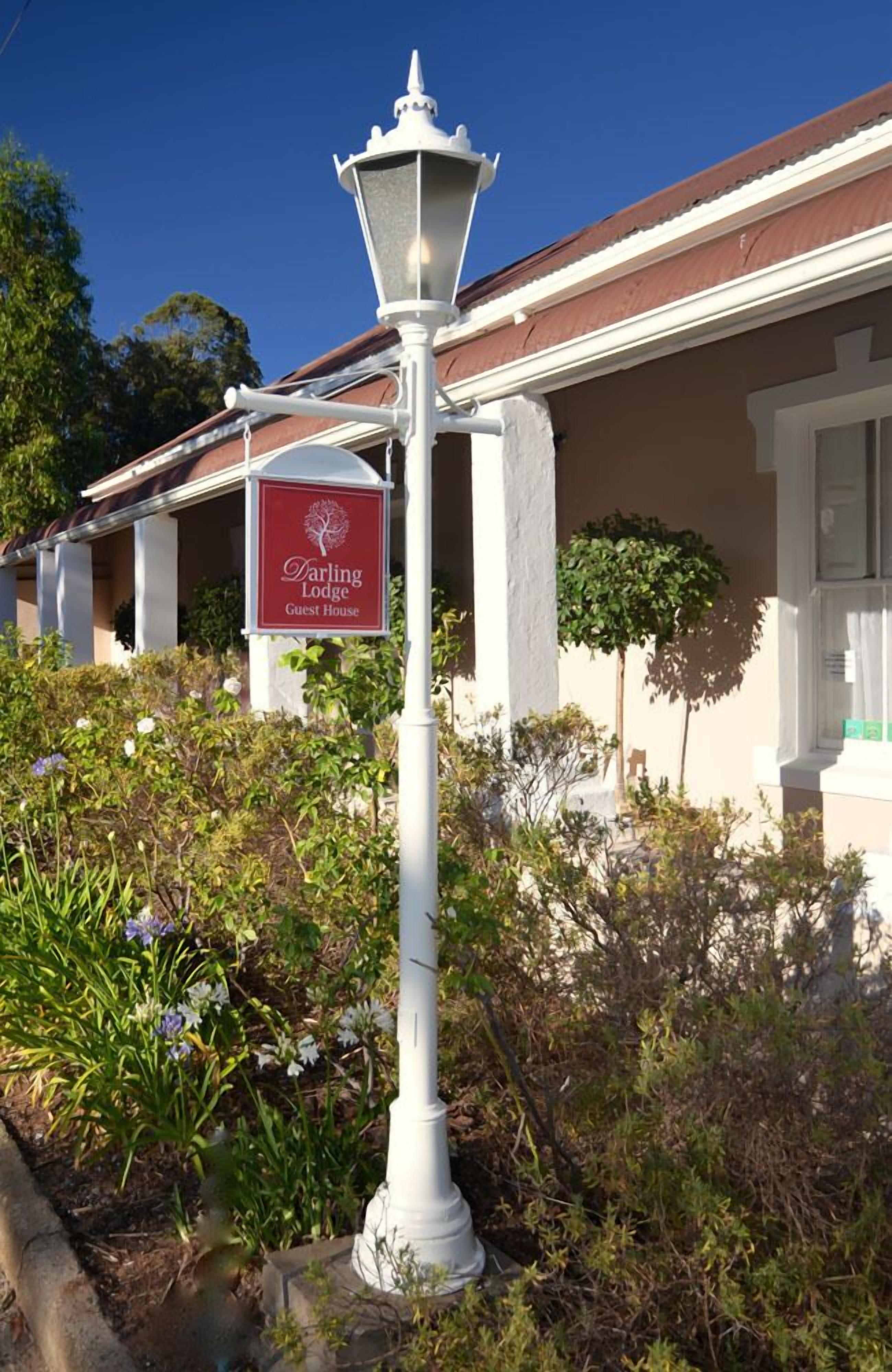 A white street lamp with a sign for Darling Lodge Guest House in front of a building with a brown roof and white trim.