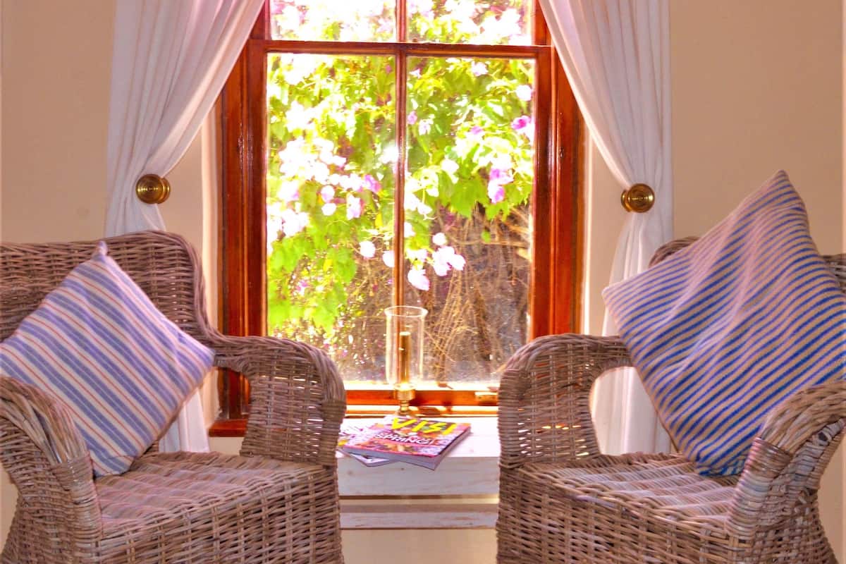 A room with two wicker chairs, each with a striped pillow, positioned in front of a window with white curtains. A vase with flowers is placed on the windowsill.