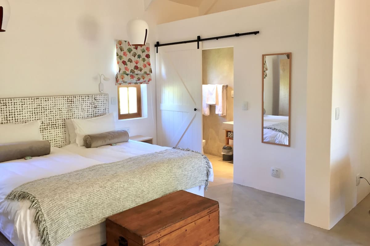 A bedroom with a bed, a wooden chest, a window with curtains, a mirror, and a door leading to another room.