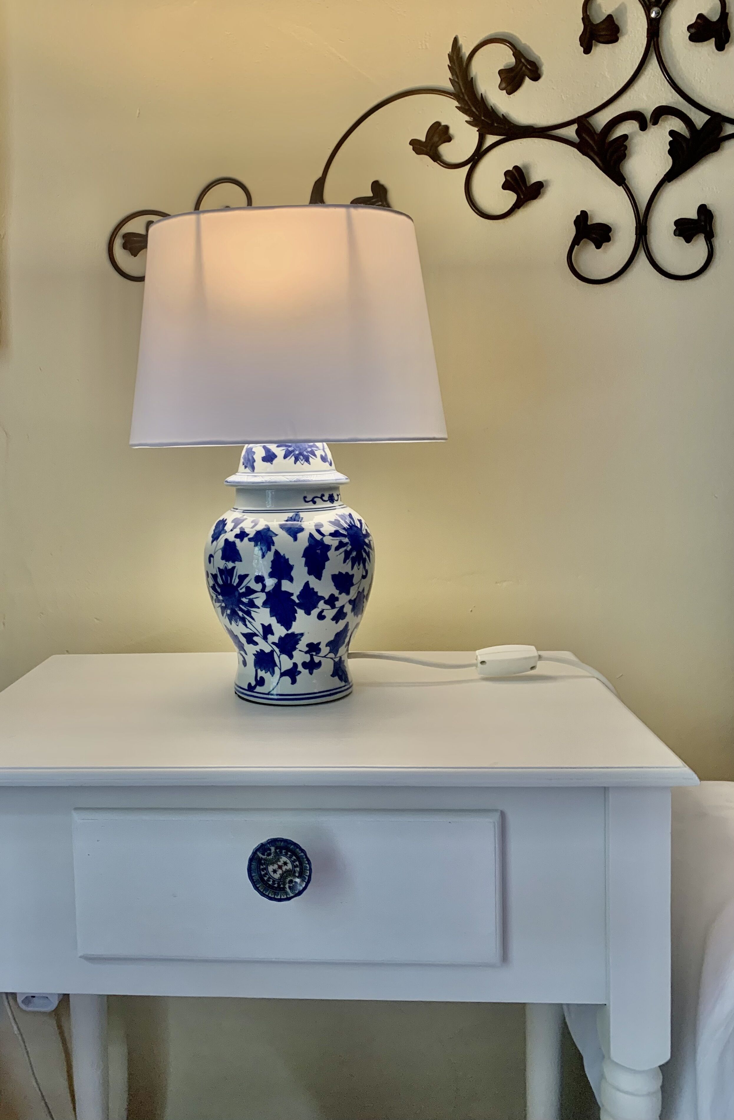 Uma mesinha de cabeceira branca com uma luminária azul e branca, um objeto decorativo na parede e uma gaveta pequena com um puxador redondo.