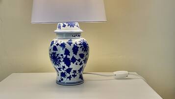 Uma mesinha de cabeceira branca com uma luminária azul e branca, um objeto decorativo na parede e uma gaveta pequena com um puxador redondo.