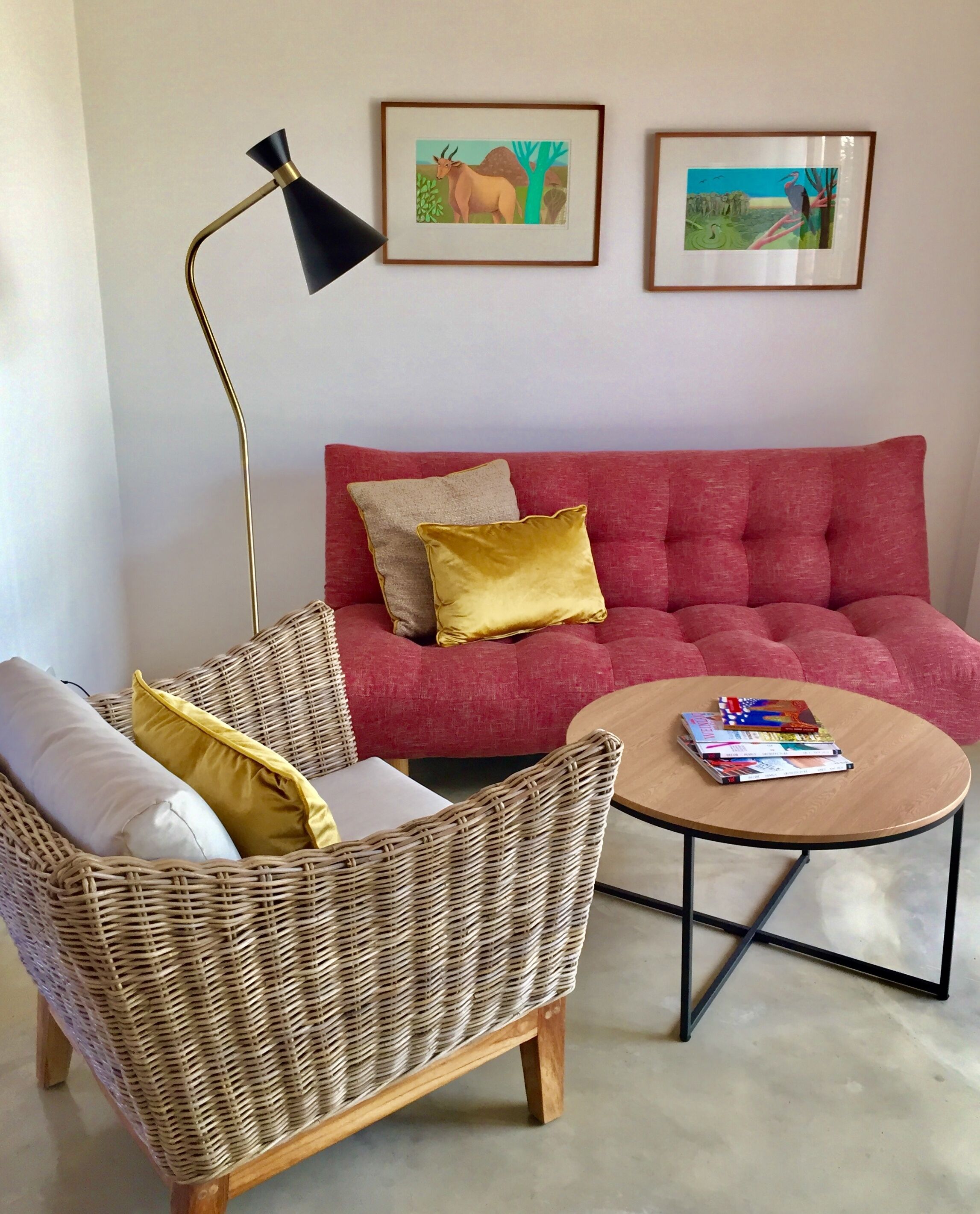 Un salon avec un canapé rouge, un fauteuil en osier, une petite table ronde sur laquelle sont posés des livres, un lampadaire et deux tableaux encadrés accrochés au mur.