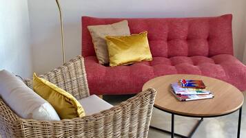 Un salon avec un canapé rouge, un fauteuil en osier, une petite table ronde sur laquelle sont posés des livres, un lampadaire et deux tableaux encadrés accrochés au mur.