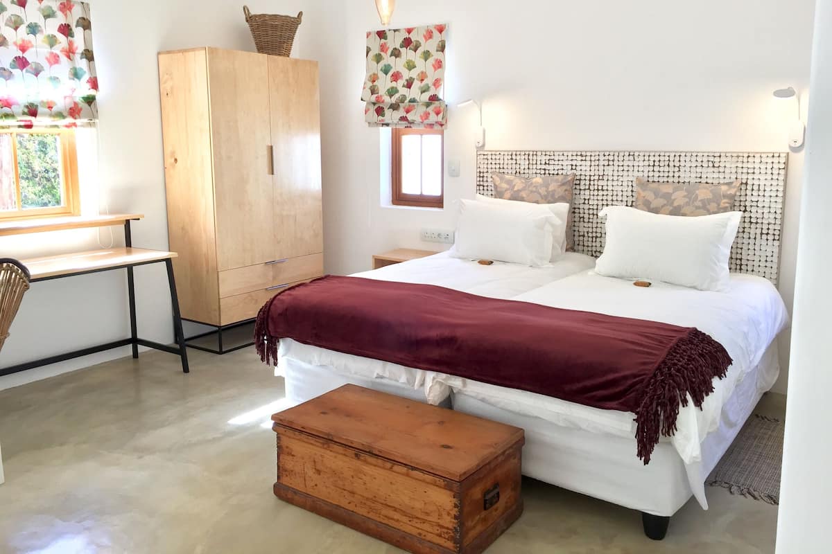 A bedroom with a wooden wardrobe, a desk, a bed with white bedding and a maroon throw, and a wooden chest of drawers.