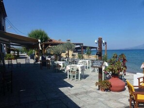 An outdoor dining area with tables and chairs, a sign indicating seafood and vegetable options, and a view of the sea. - Paris Beach (Samos)