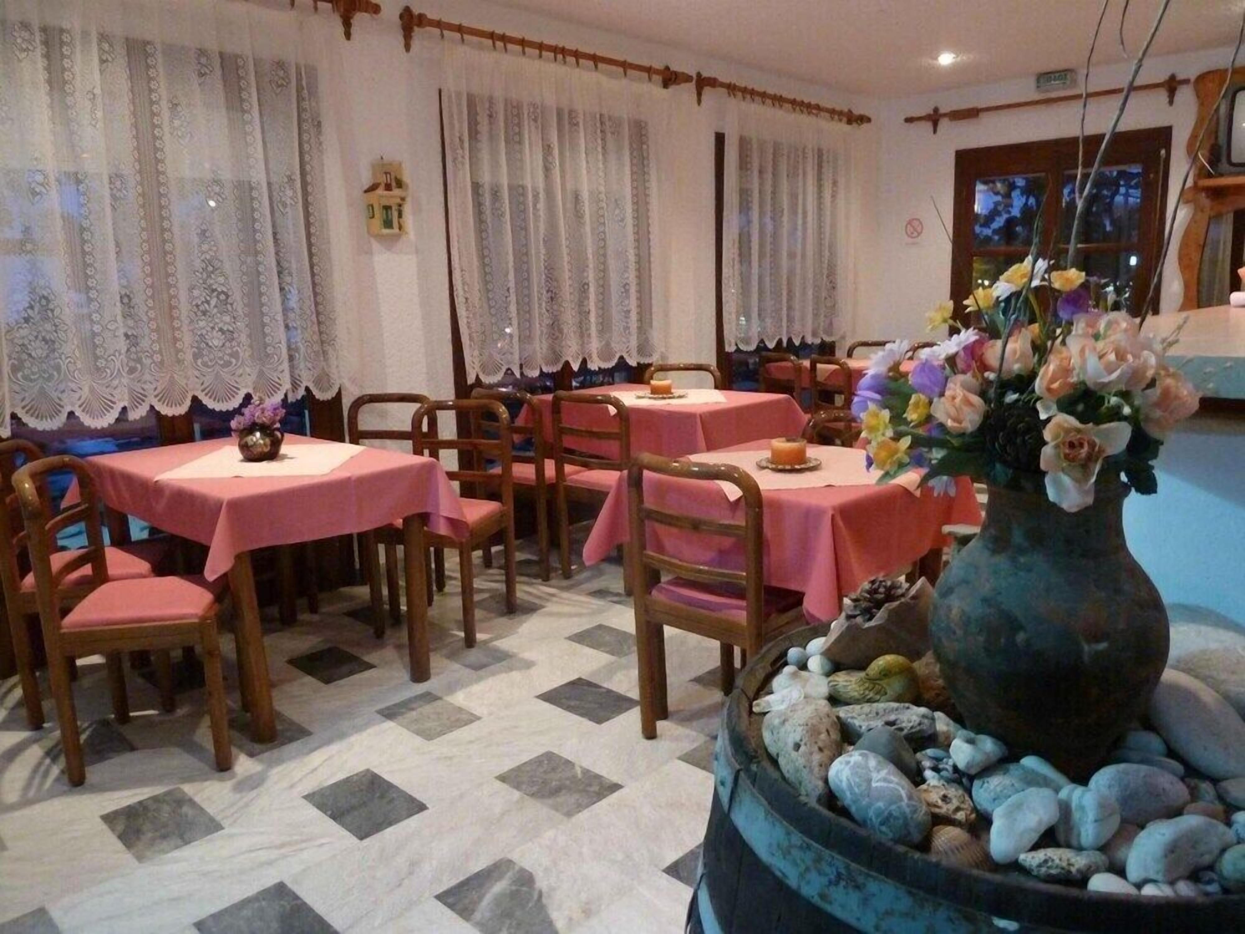 A restaurant with tables covered in pink tablecloths, wooden chairs, lace curtains, and a decorative vase with flowers.