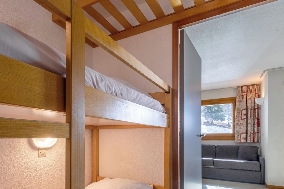 A bunk bed room with a wooden frame, a grey sofa, and a window with curtains.