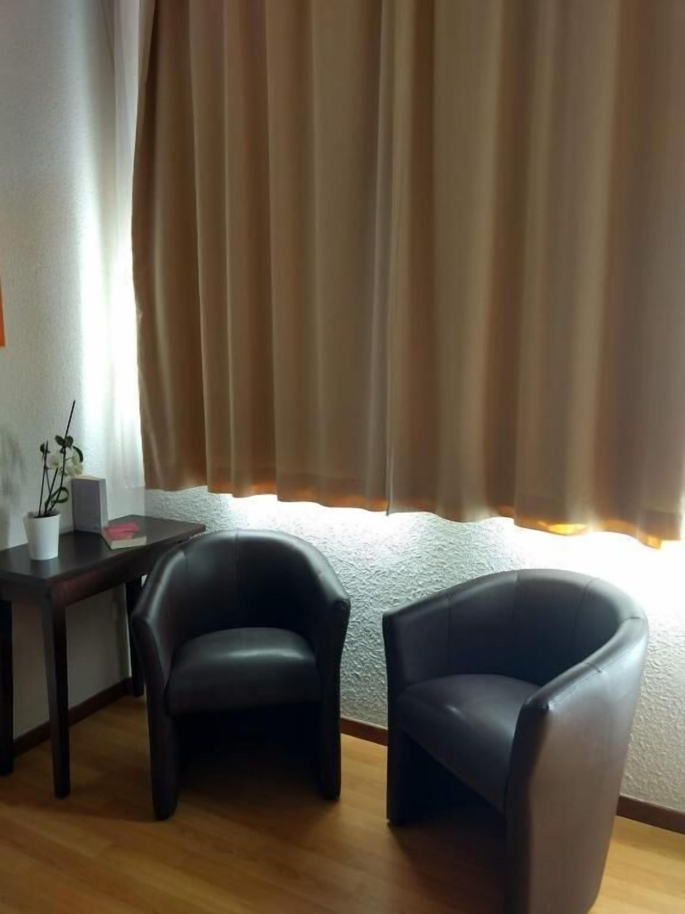 A room with two black leather chairs, a wooden table with a potted plant and a book, and a window with beige curtains.