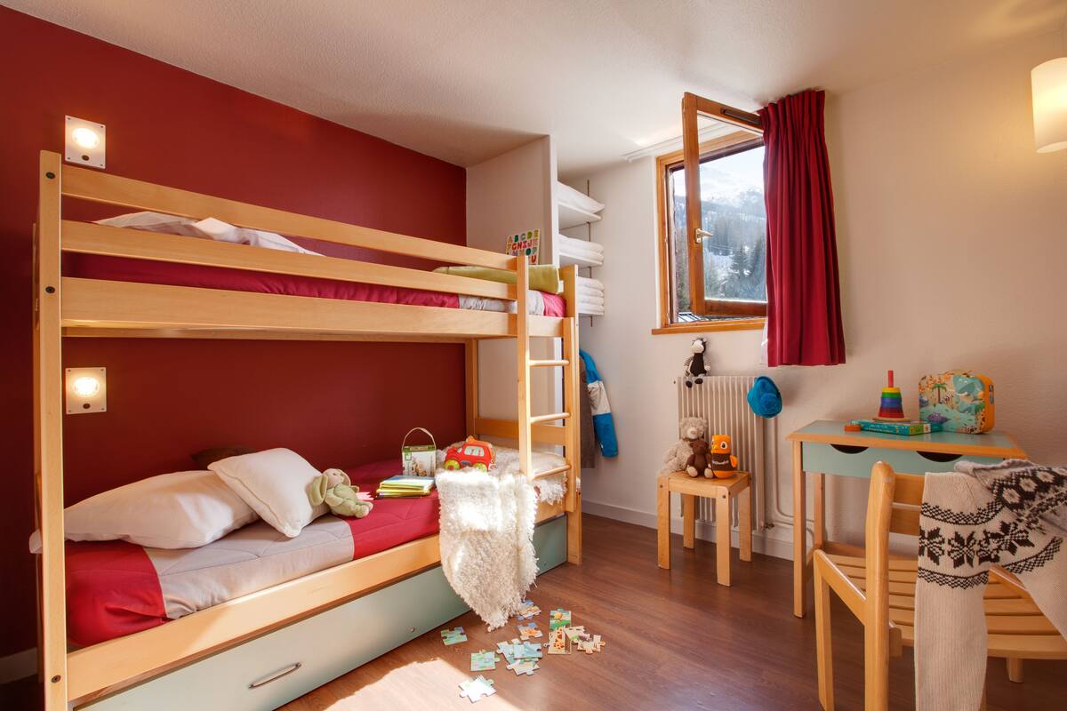 A bunk bed room with a desk and chair, a window with curtains, and a radiator.