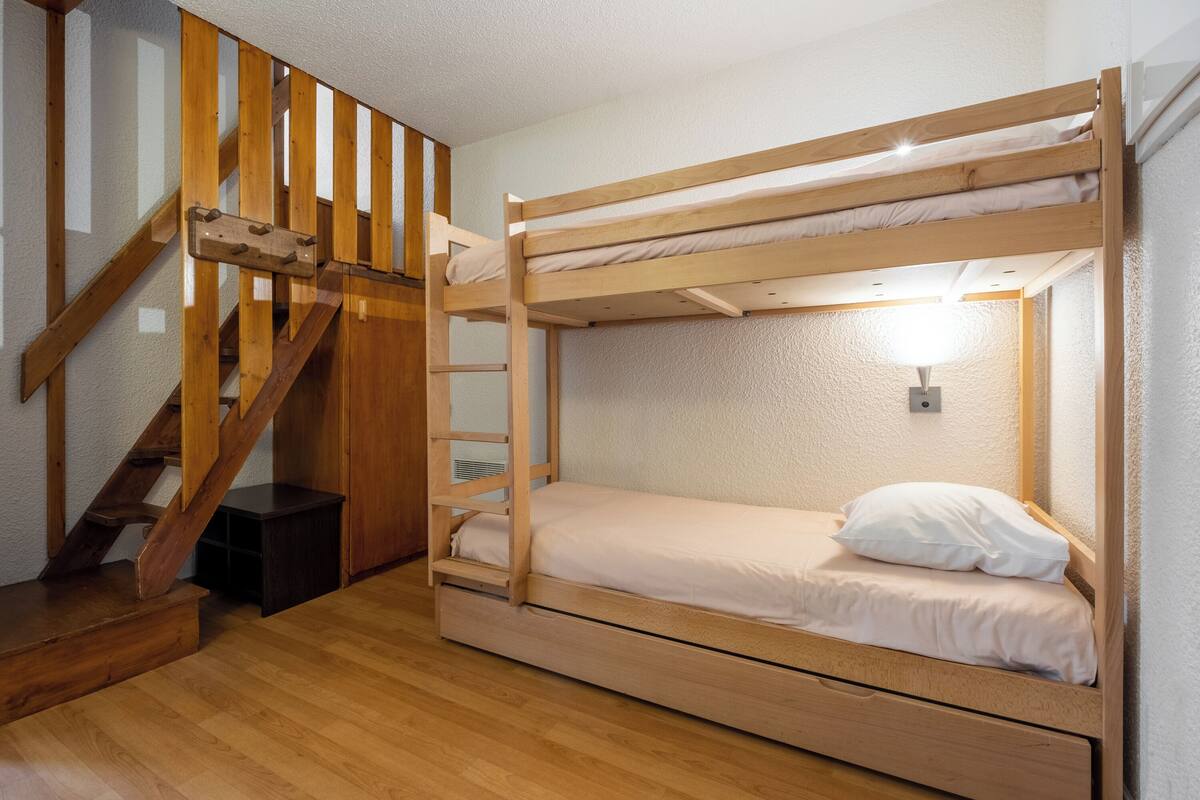 A bunk bed with a wooden ladder, a small desk, and a wall-mounted light fixture.
