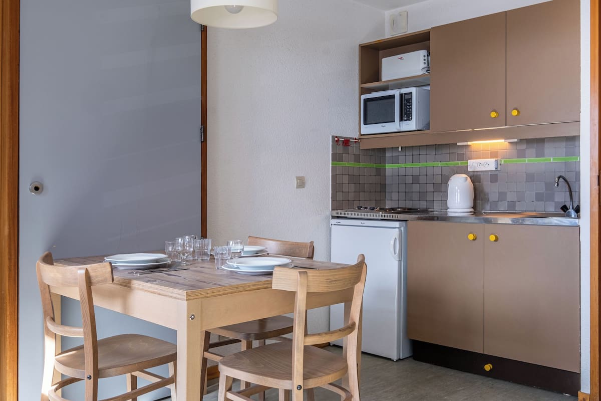 A compact kitchen with a microwave, kettle, and a small refrigerator. There is a wooden dining table with chairs and a few plates and glasses set.