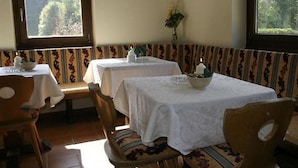 A dining area with two tables, each covered with a white tablecloth. There are wooden chairs with patterned cushions. A vase with flowers is on the wall, and a window provides a view of trees. - Landhotel Traudl (Saalbach-Hinterglemm)