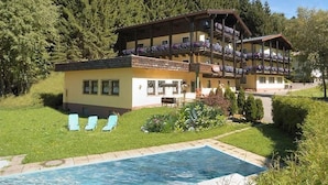 A two-story building with a pool, surrounded by greenery and trees. - Landhotel Traudl (Saalbach-Hinterglemm)