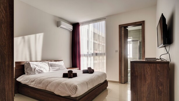 A modern hotel room with a large bed, a wooden headboard, a TV mounted on the wall, and a large window with sheer curtains. - The Village Apartments (St. Paul's Bay)
