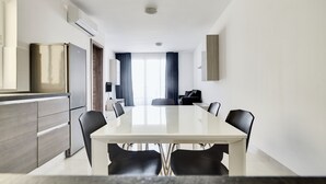 A modern hotel room with a dining area featuring a large table and chairs, a kitchen area with a refrigerator, and a seating area with a sofa and chairs. - The Village Apartments (St. Paul's Bay)