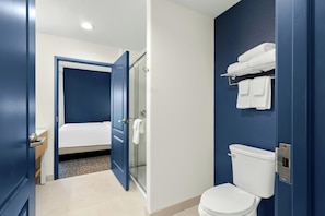 A hotel bathroom with a white toilet, a glass shower enclosure, and a bed in the adjacent room. - Spark by Hilton Missouri City (Missouri City)