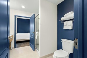 A hotel bathroom with a white toilet, a glass shower enclosure, and a bed in the adjacent room. - Spark by Hilton Missouri City (Missouri City)