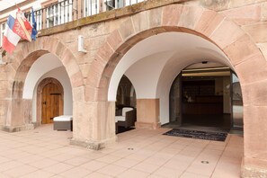 A stone building with arched entrances, a wooden door, and a bench. Flags are visible above the entrance. - Val-vignes Haut-koenigsbourg, The Originals Relais (Saint-Hippolyte)