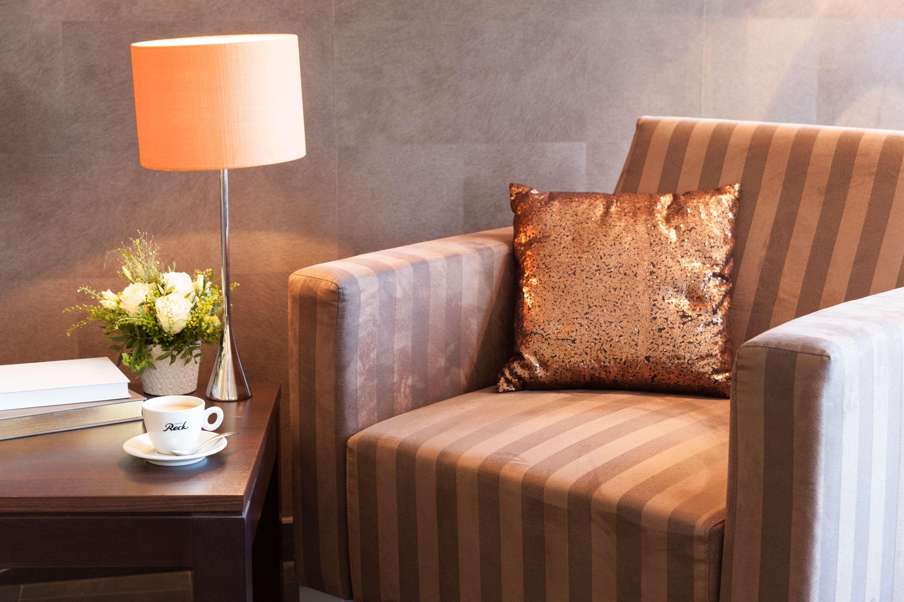 Una habitación de hotel con un sillón a rayas, un cojín decorativo, una mesita de noche con una lámpara, una taza y un florero con flores.