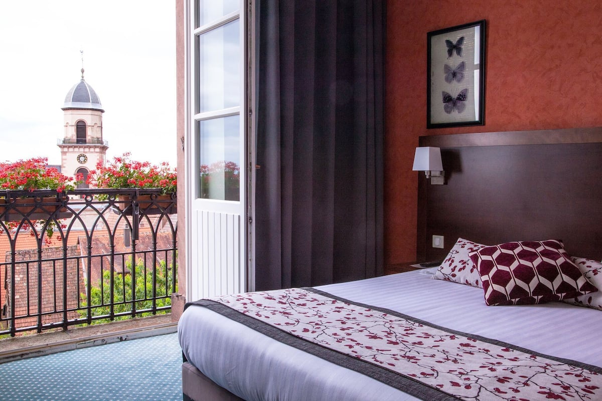 Une chambre d’hôtel avec un lit, une fenêtre avec des rideaux, un tableau représentant un papillon encadré, et une vue sur une tour et des fleurs à l’extérieur.