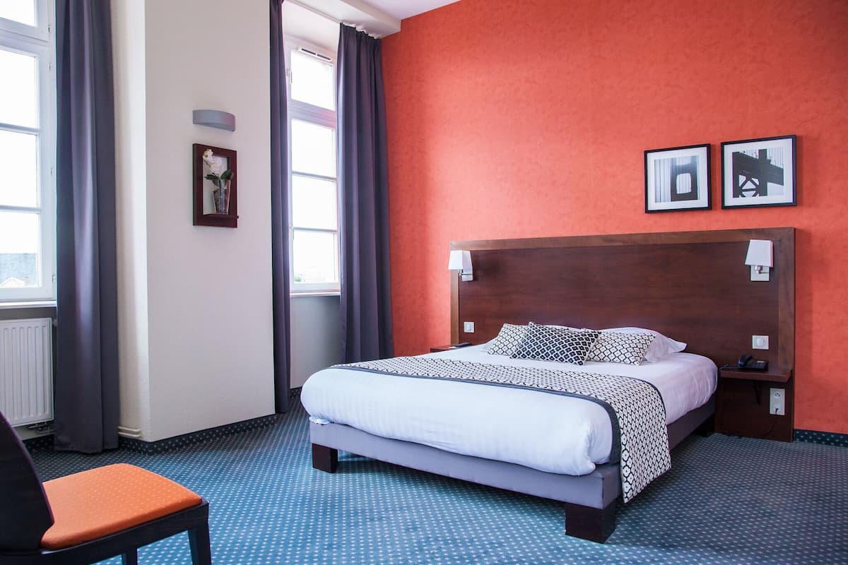 Une chambre d’hôtel avec un grand lit, une tête de lit en bois, une table de chevet, une chaise et une fenêtre avec des rideaux.