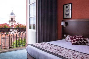 Une chambre d’hôtel avec un lit, une fenêtre avec des rideaux, un tableau représentant un papillon encadré, et une vue sur une tour et des fleurs à l’extérieur.