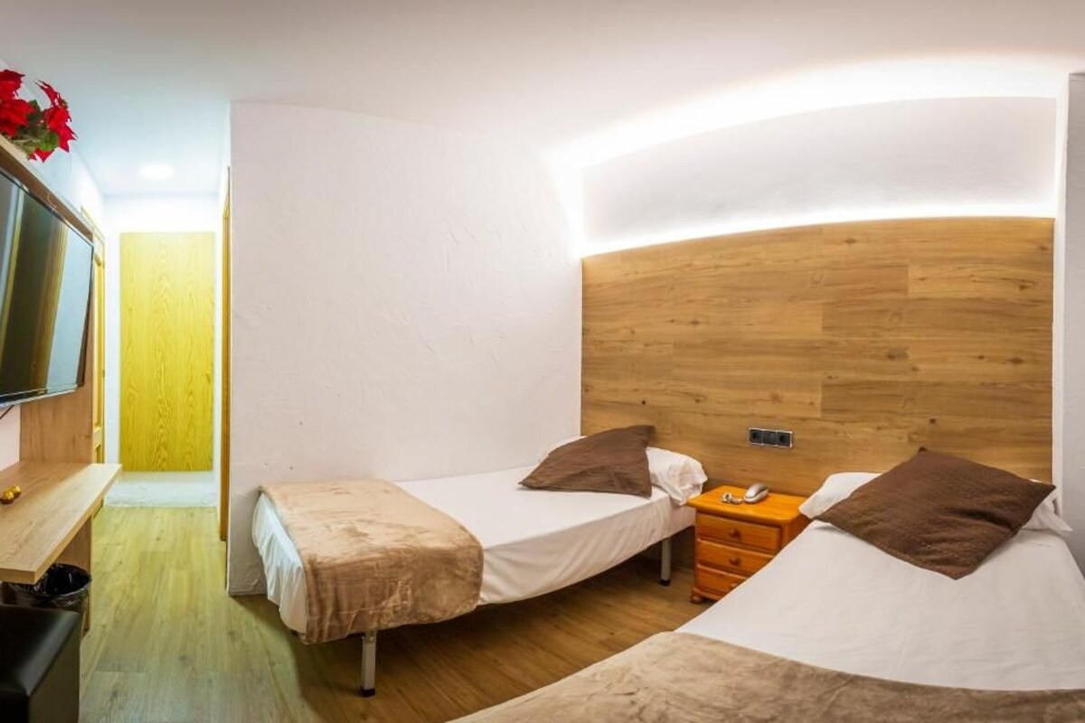 A compact hotel room with two single beds, a wooden headboard, a small desk, and a television.