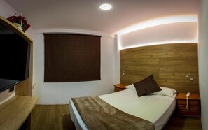 Un dormitorio con cabecera de madera, cama con colchón blanco y manta marrón, mesita de noche con lámpara y televisor instalado en la pared.