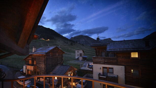 Vista desde un balcón sobre un pueblo rural al anochecer, con casas de madera y una montaña en el fondo.