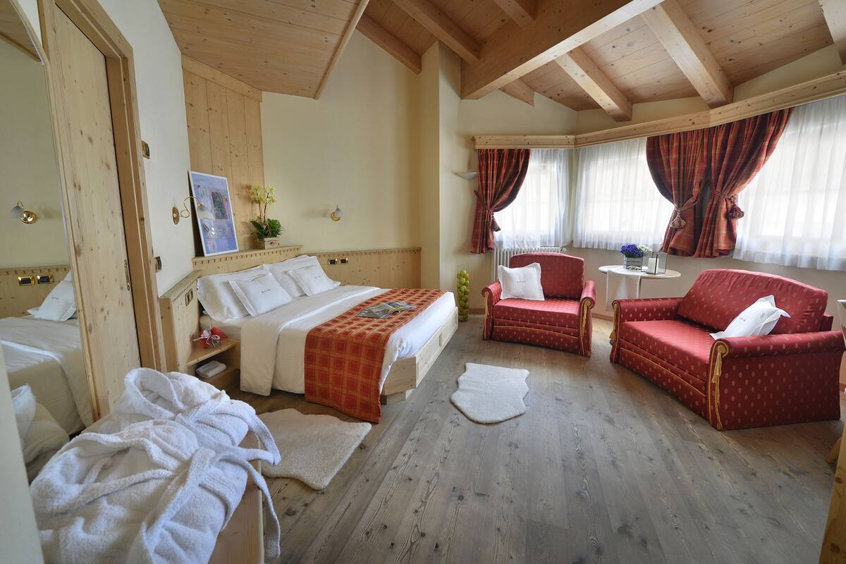 A hotel room with a wooden ceiling, a bed with a red blanket, two red armchairs, and a small table with a vase of flowers.