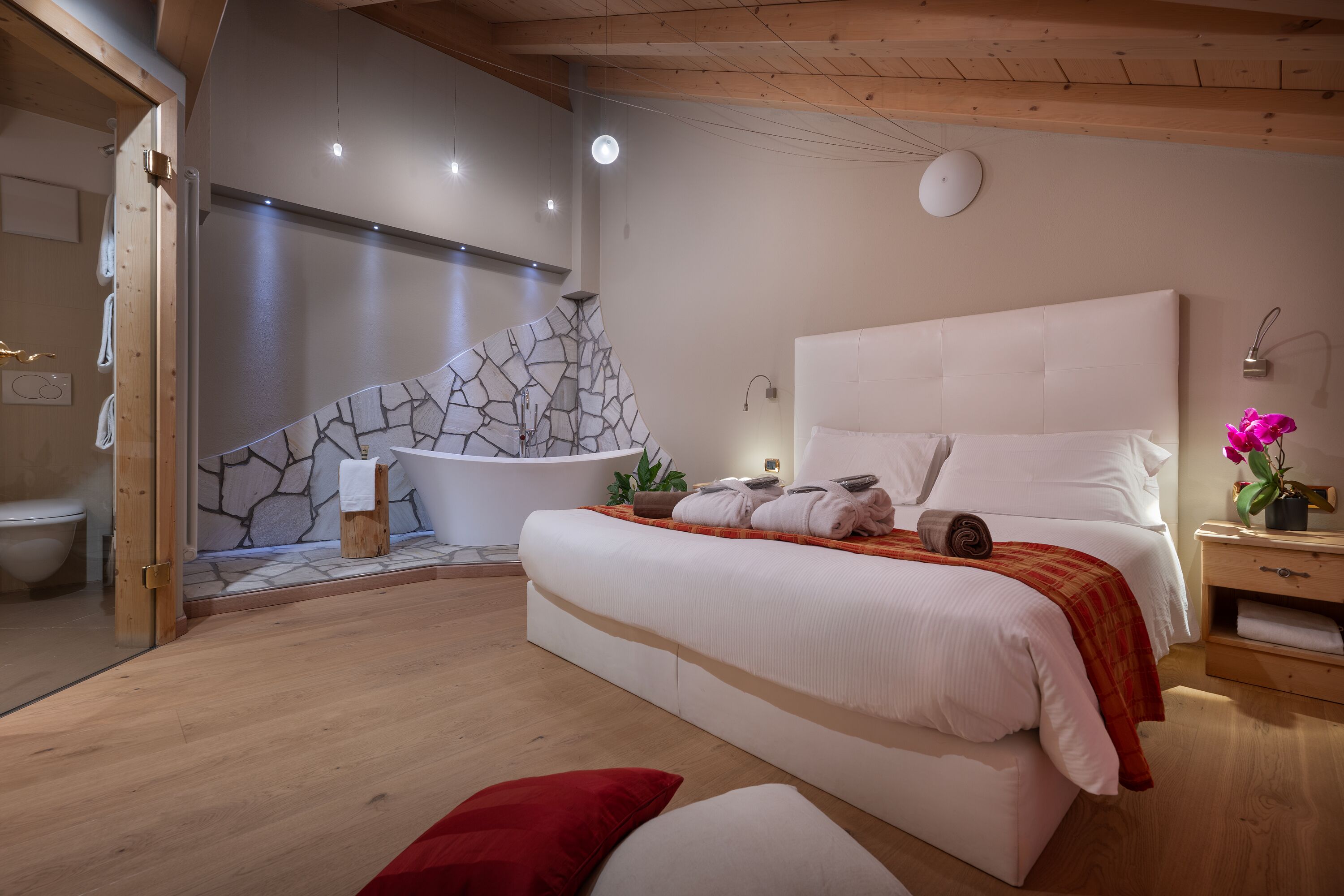 Une chambre moderne avec un mur en pierre, un grand lit, un plancher en bois et une salle de bain équipée de toilettes et d’un lavabo.