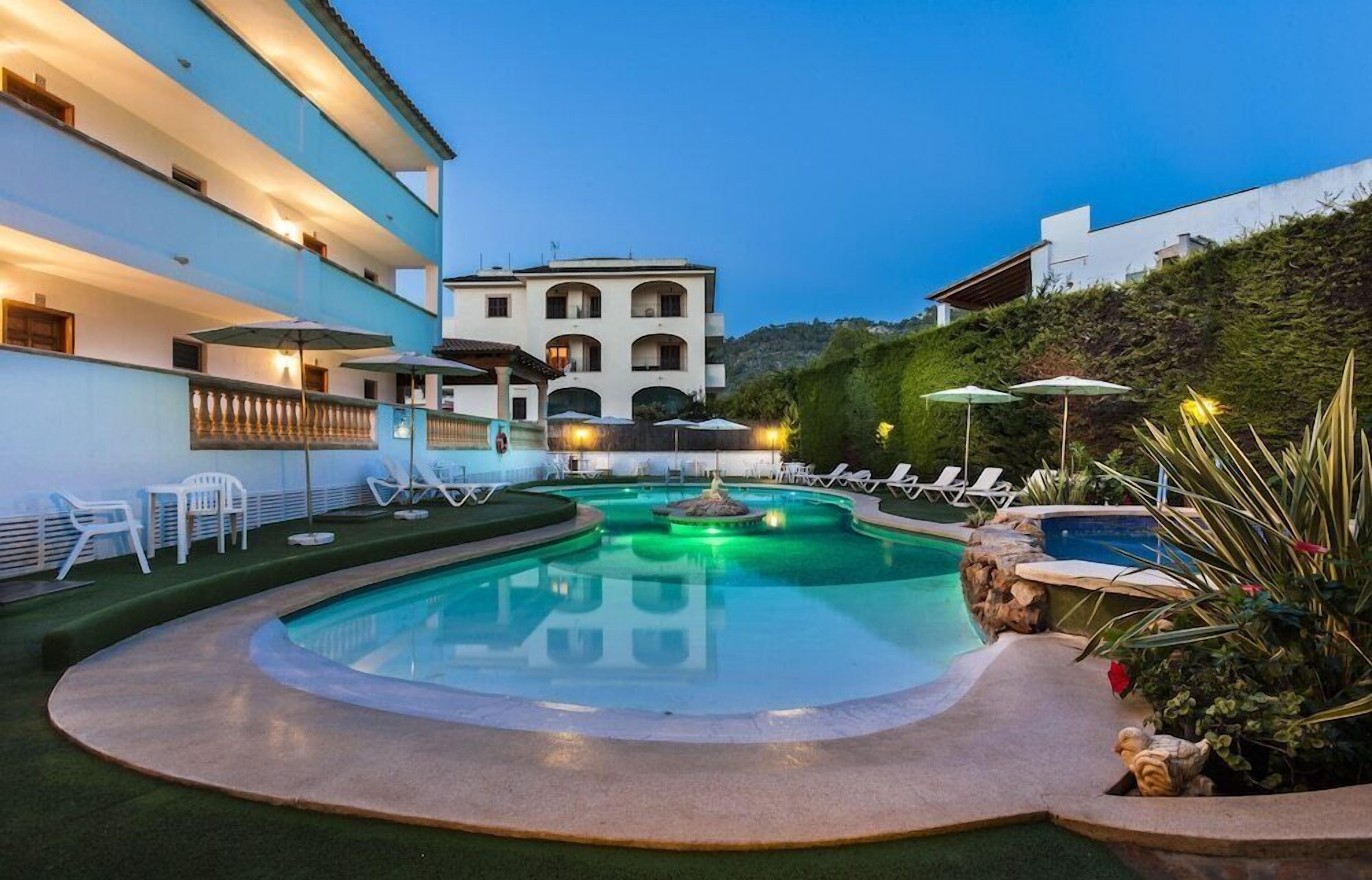 Una piscina con sillas de descanso y sombrillas, rodeada de edificios y zonas ajardinadas.