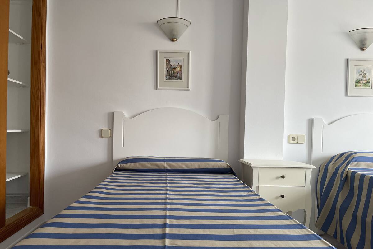 A single bed with a striped bedspread, a bedside table, a nightstand, and a wall-mounted light fixture.