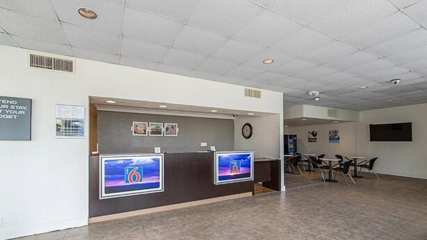 A reception area with a counter, two monitors, a clock, and a seating area with tables and chairs. - Concorde Inn Suites Pekin (Pekin)