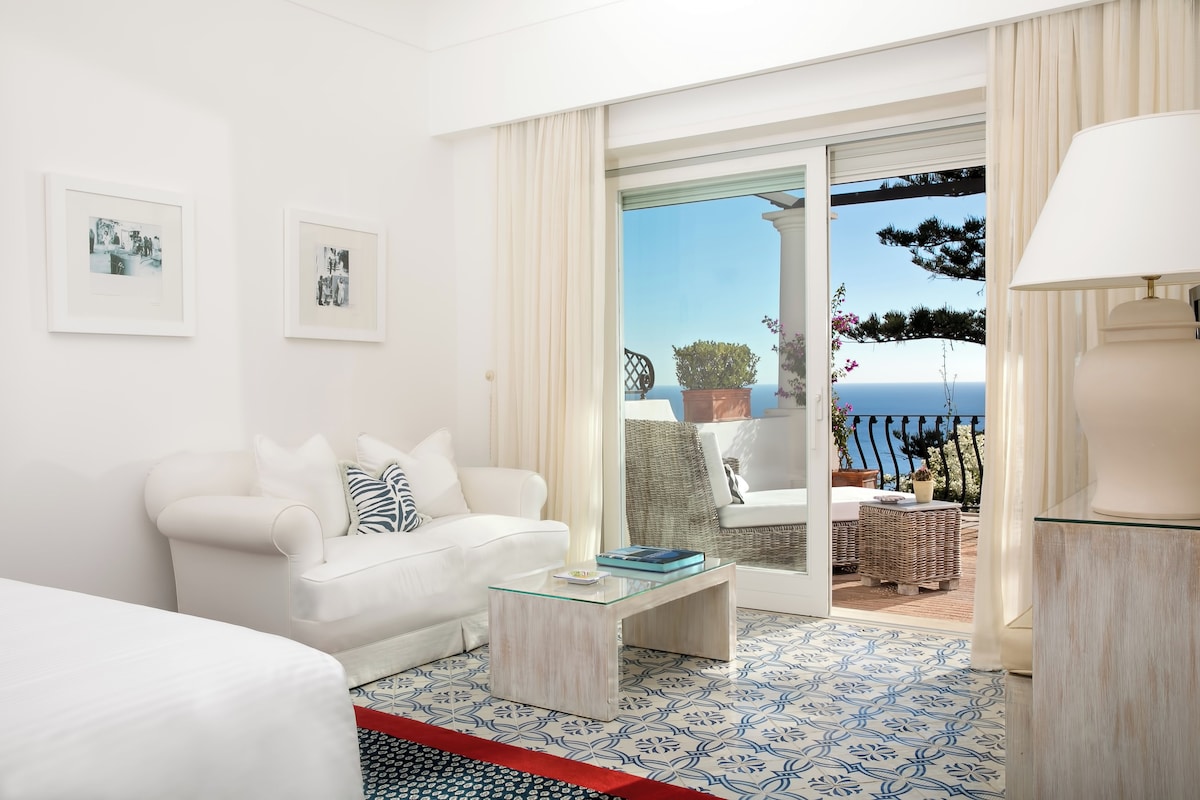 A modern living room with a white sofa, a glass coffee table, and a view of the sea through a sliding glass door.