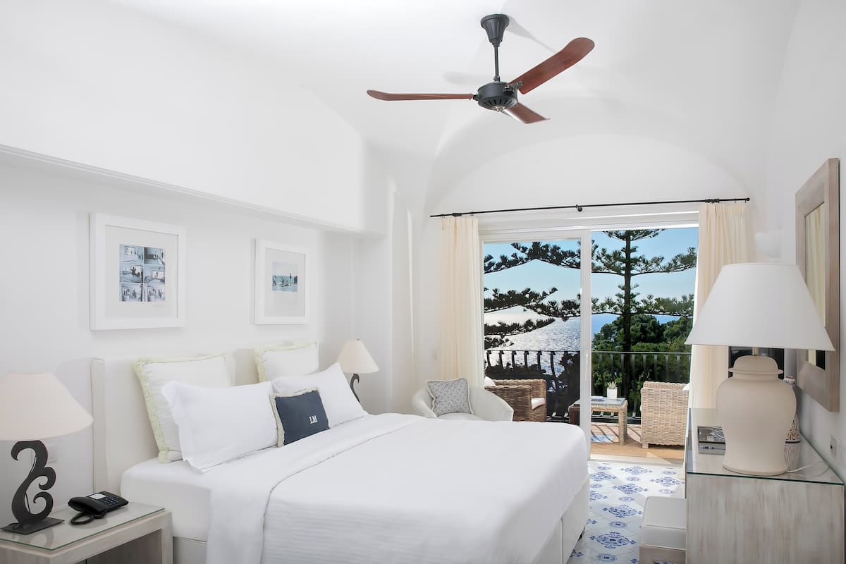 A hotel room with a bed, two bedside tables with lamps, a ceiling fan, and a balcony with outdoor furniture.