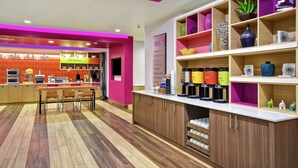 A modern cafe with a vibrant pink and orange color scheme, featuring a counter with coffee machines, a seating area, and shelves with decorative items. - Home2 Suites by Hilton Oklahoma City Airport (Oklahoma City)