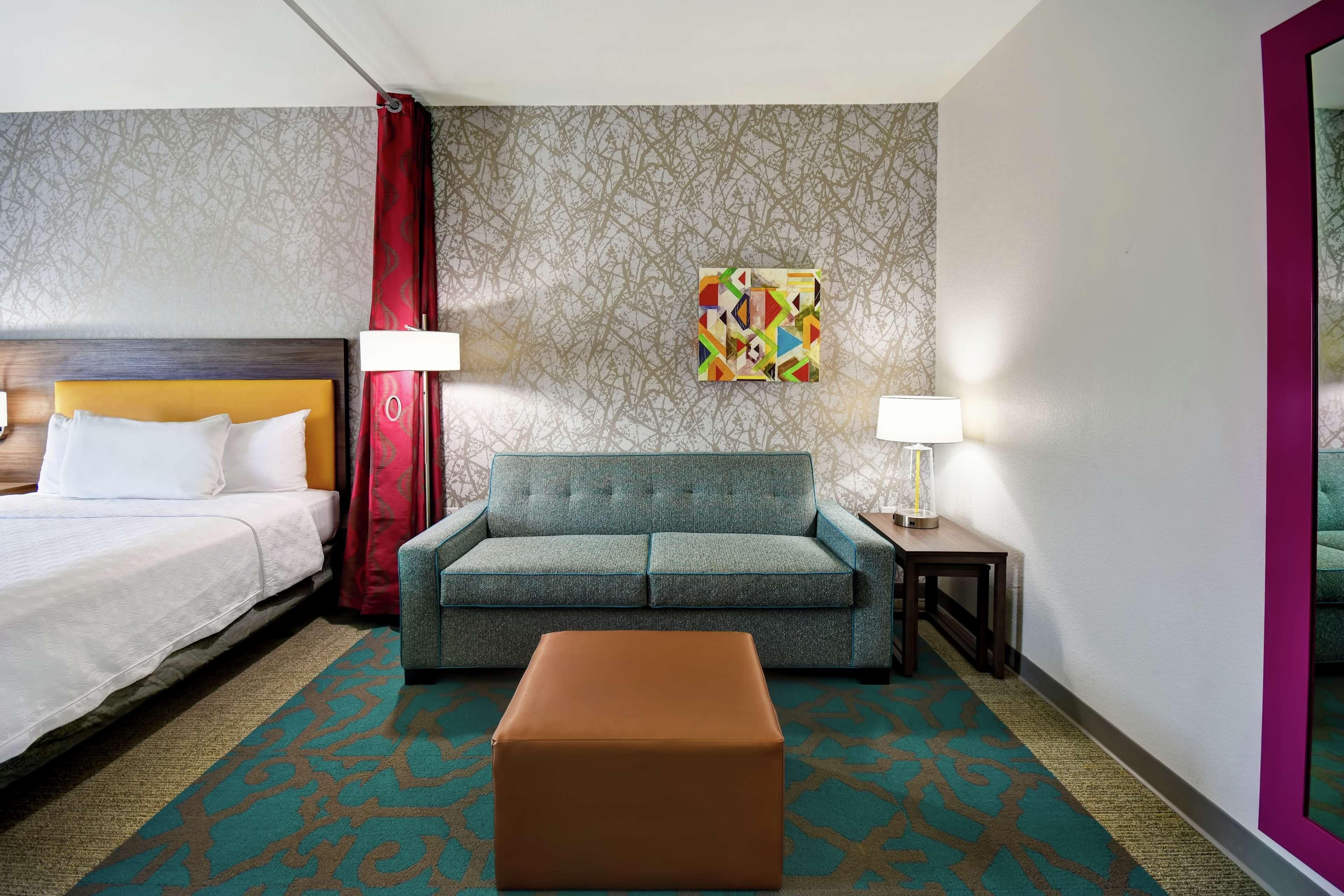 Een hotelkamer met een bed, een slaapbank, een voetenbankje, een nachtkastje, een lamp, een schilderij en behang met een patroon.