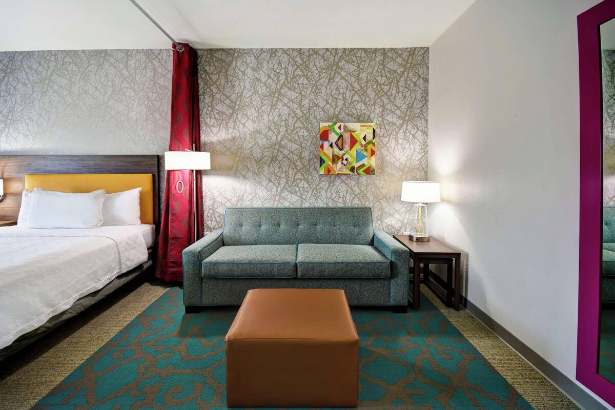 Ein Hotelzimmer mit Bett, Schlafsofa, Hocker, Nachttisch, Lampe, einem Gemälde und Wandtapete mit Muster.