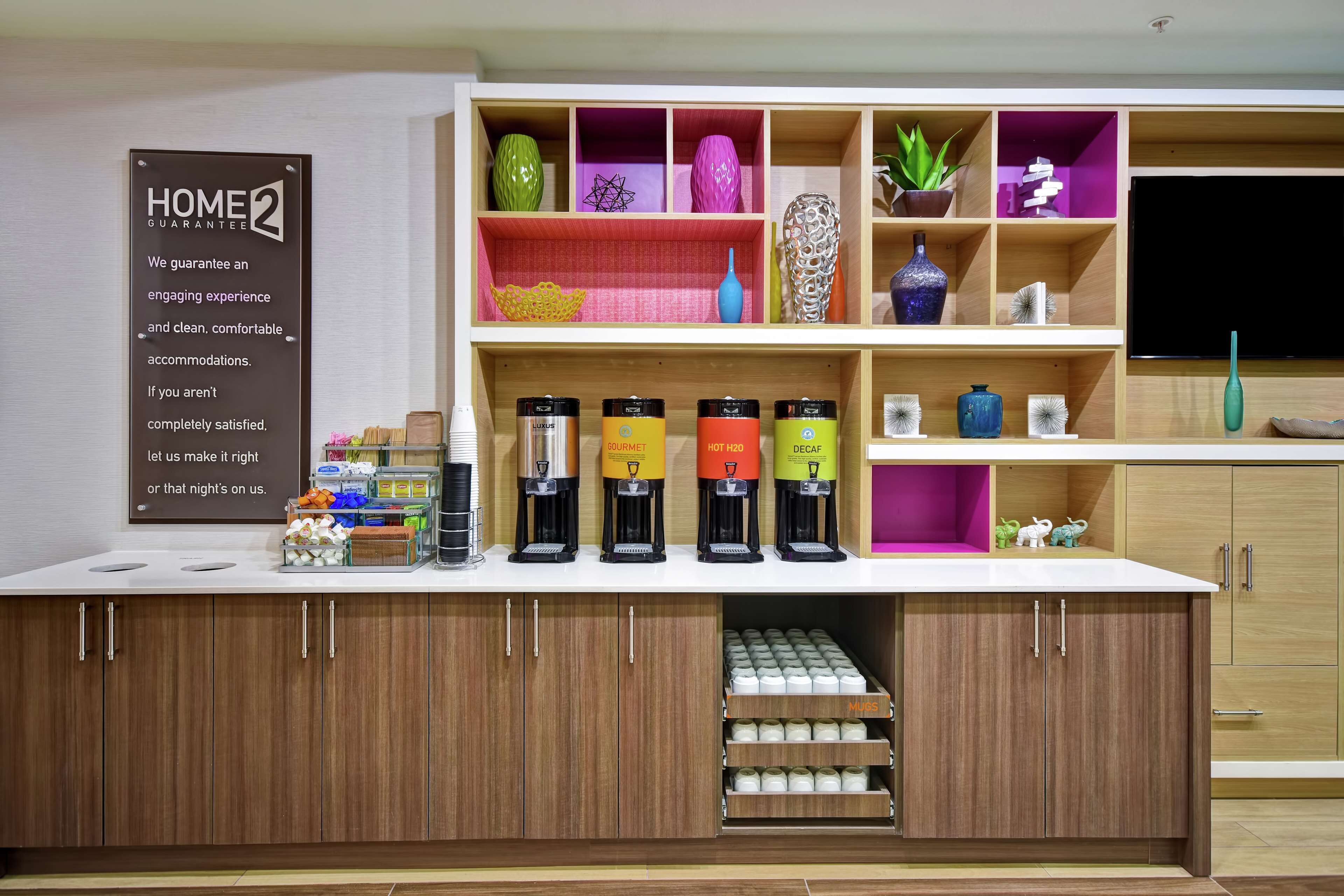 A hotel reception area with a coffee station, a 'Home 2 Guarantee' sign, and decorative shelves with plants and vases.