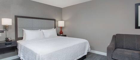 A hotel room with a large bed, two bedside tables with lamps, a grey sofa, and a carpeted floor.