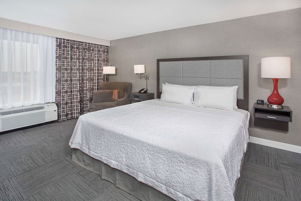 A hotel room with a large bed, a chair, a nightstand with a lamp, a wall-mounted phone, and a window with curtains.