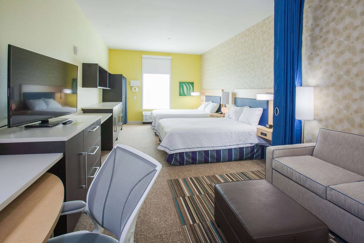 A hotel room with a desk, two beds, a sofa, a TV, and a bathroom.