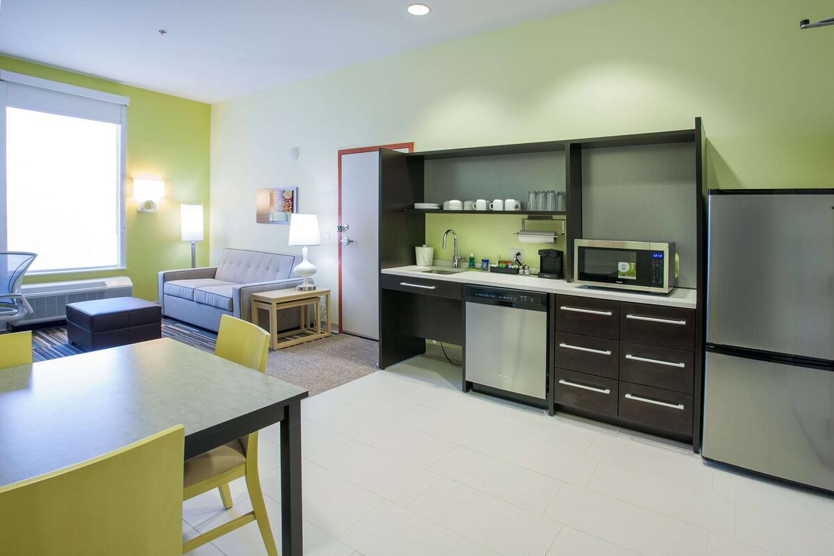 A compact kitchen with a microwave, refrigerator, and dishwasher. There is a dining table with chairs and a sofa in the adjacent living area.