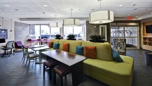 A modern lounge area with a green sofa, wooden table, and chairs, featuring ceiling lights and large windows. - Home2 Suites by Hilton Brownsville (Brownsville)